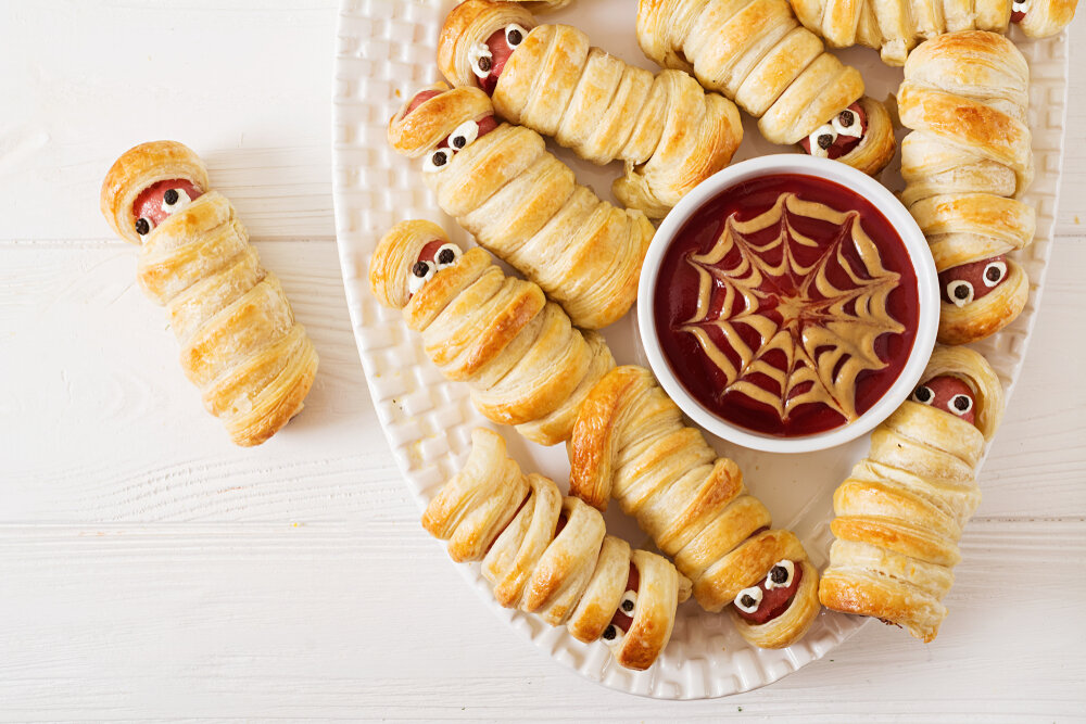 Golden pastry-wrapped mummy dogs with edible eyes and a bowl of dipping sauce